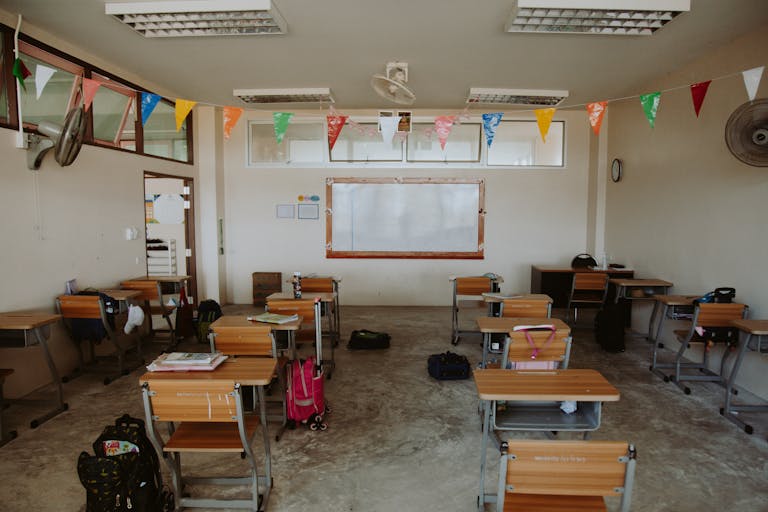An empty classroom with desks arranged neatly and colorful flags hanging from the ceiling, creating a bright and hopeful atmosphere.