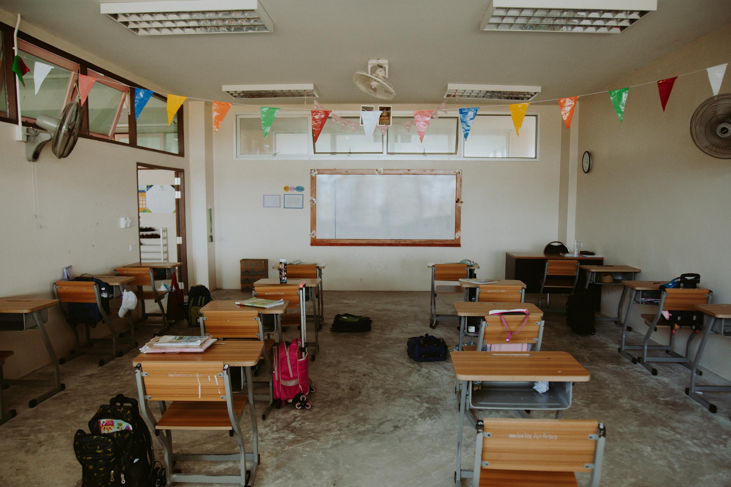 An empty classroom with desks arranged neatly and colorful flags hanging from the ceiling, creating a bright and hopeful atmosphere.
